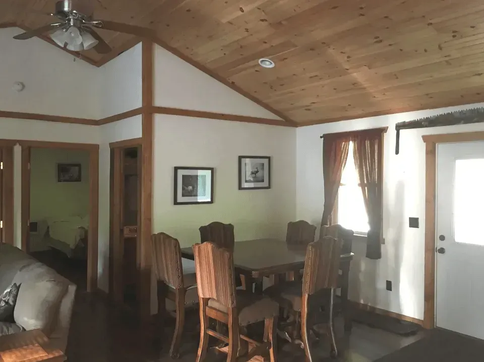 A dining area with a wooden table and chairs, set beneath a vaulted wood-paneled ceiling next to a window and doorway.