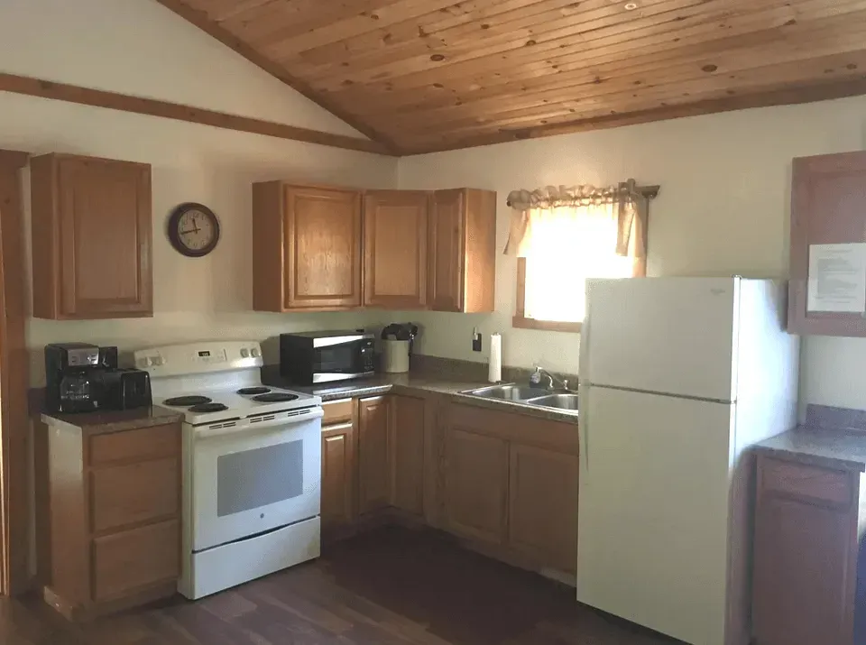 A modern kitchen with off-white cabinets, a black range hood, a marble-topped island, and wooden chairs in the foreground.