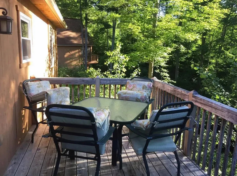 A glass-topped table with four chairs on a wooden deck overlooking a lush green forest.