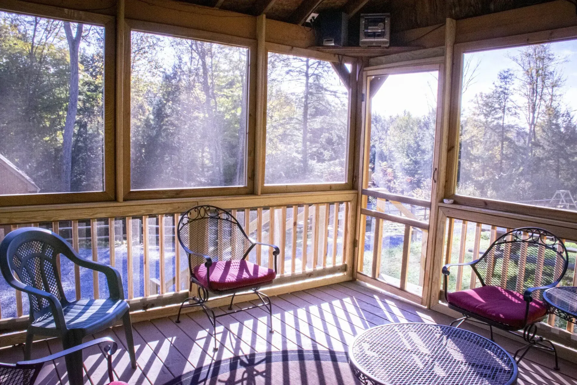 A screened-in wooden porch with three chairs and a small table overlooking a wooded area.