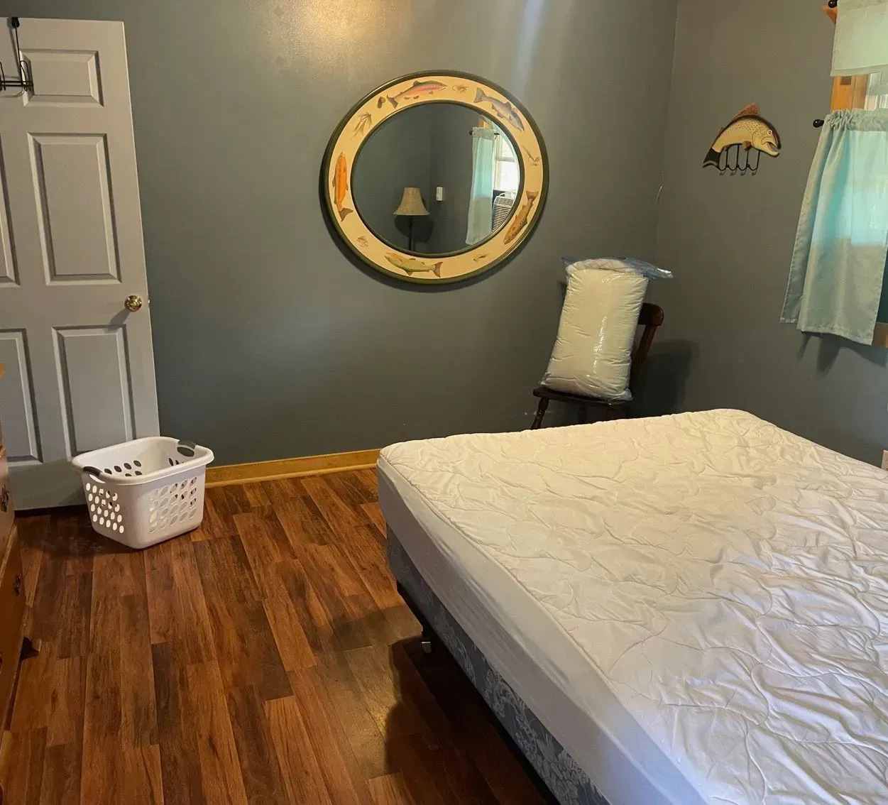 A bedroom with grey walls, hardwood flooring, a mattress on a base, a wall-mounted oval mirror, and a laundry basket.