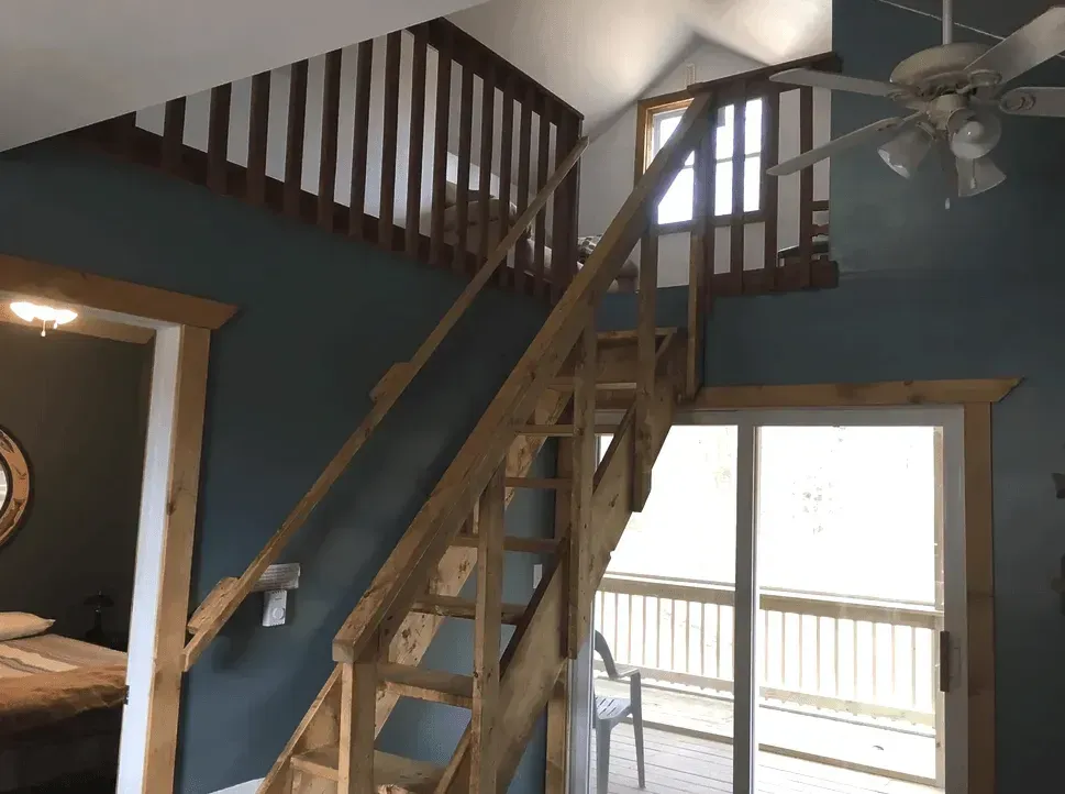 A wooden staircase leads to an upper loft in a room with dark blue walls, a ceiling fan, and sliding glass doors.
