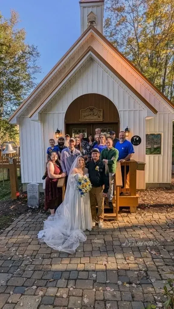 A wedding party stands together on a brick patio in front of a white wooden chapel on a sunny day.
