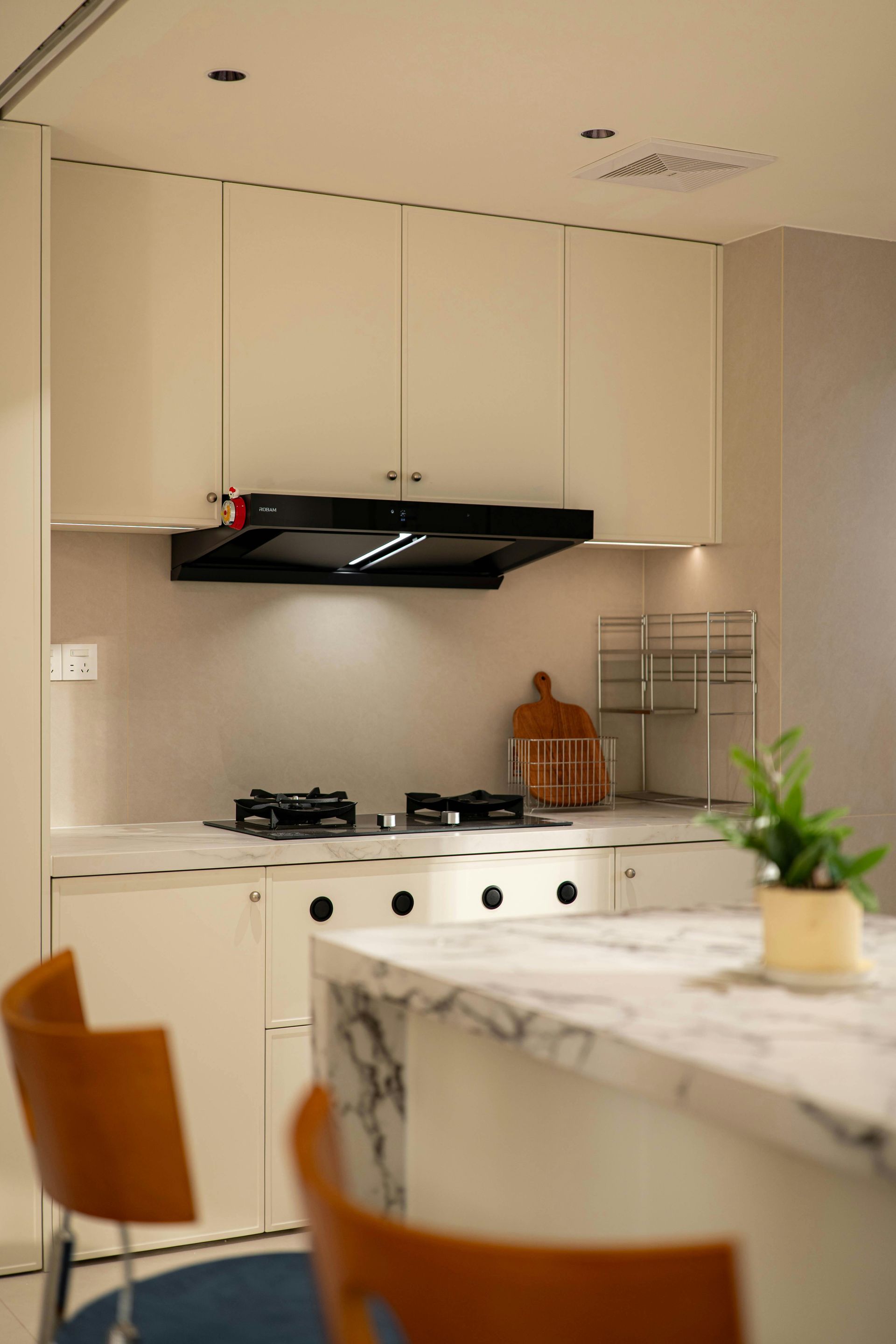 A modern kitchen with off-white cabinets, a black range hood, a marble-topped island, and wooden chairs in the foreground.