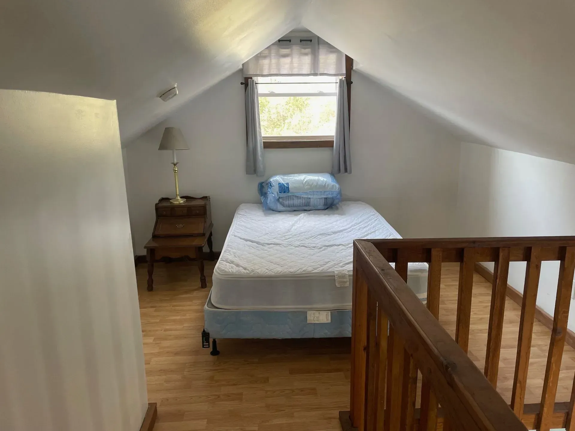 An attic bedroom with a single bed, a small wooden nightstand with a lamp, and a window under a slanted ceiling.