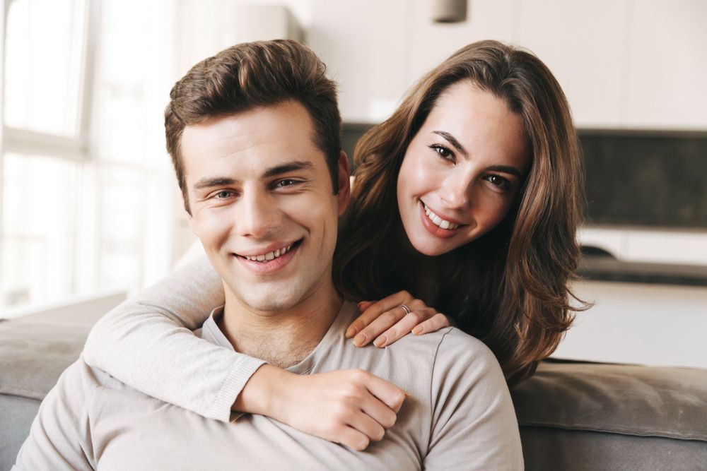 Woman hugs man from behind; both smile, indoors.