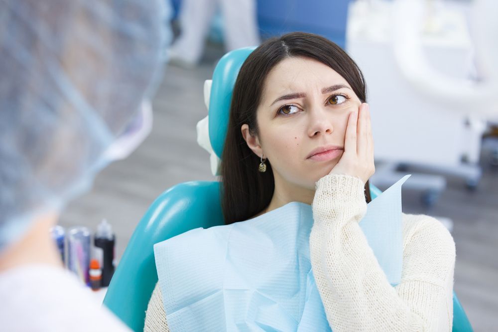 Woman in dental chair, holding jaw, looking distressed, doctor in background.