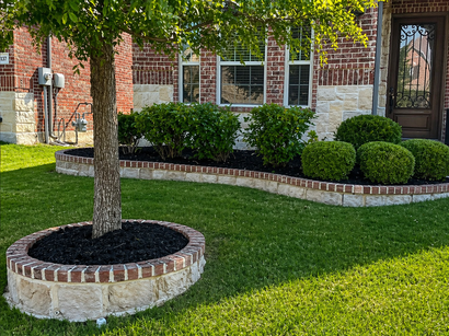 Front yard with brick house, manicured shrubs, and a tree in a stone planter on a green lawn.