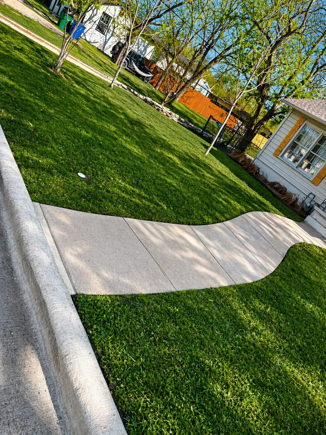 Curved concrete sidewalk beside a green lawn, trees, and a house on a sunny day.