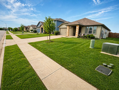 Suburban street with single-story homes, green lawns, and a sidewalk under a clear sky