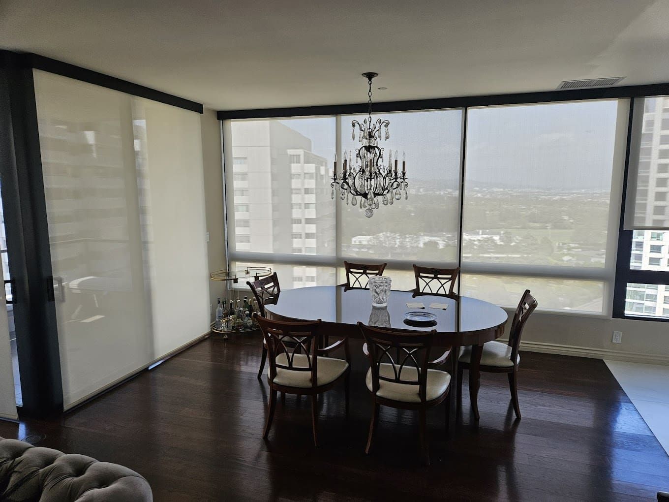 Dining room with oval table, chairs, chandelier, and city view through windows.