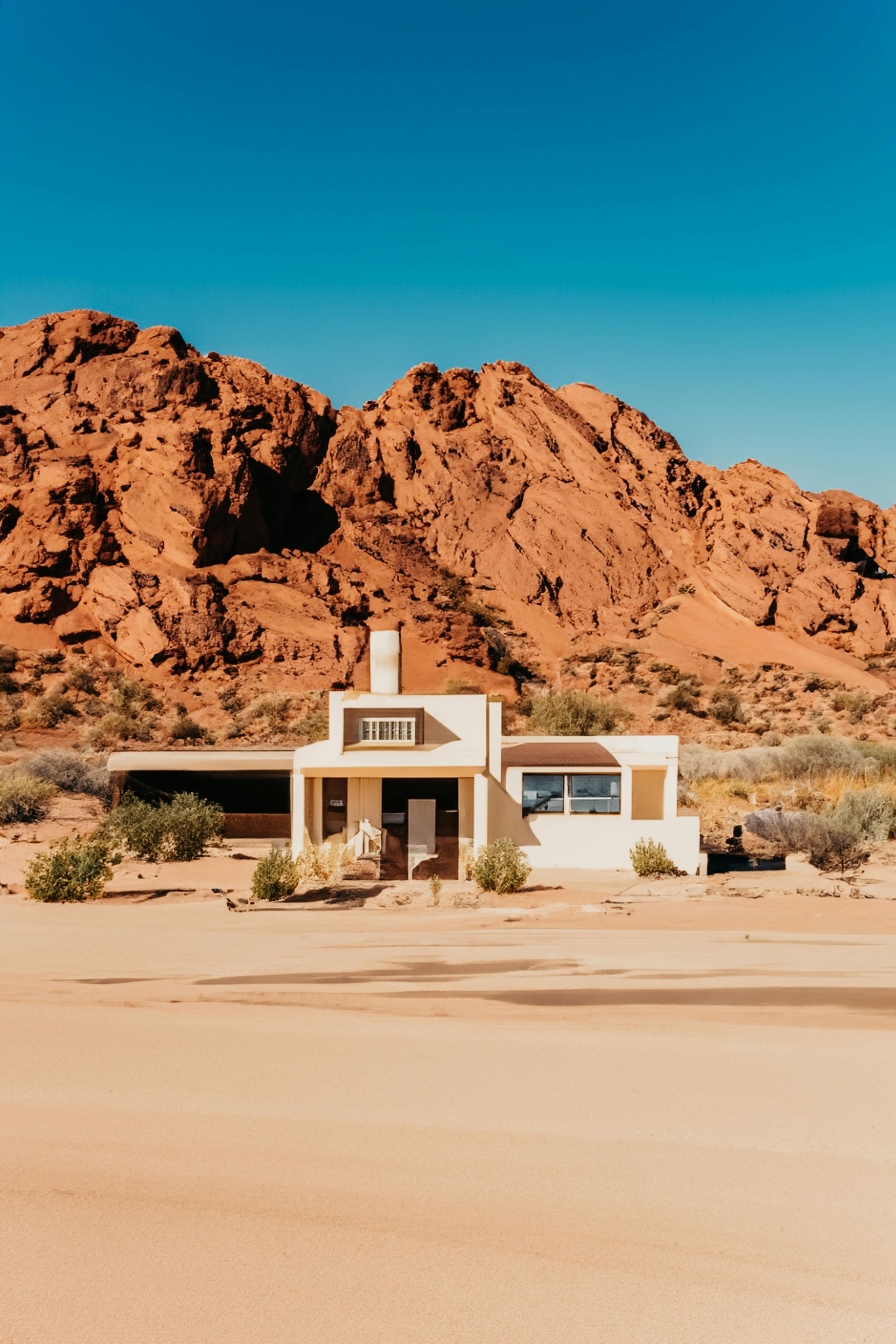 Desert landscape with a small beige house against reddish-brown rocky mountains and a bright blue sky.