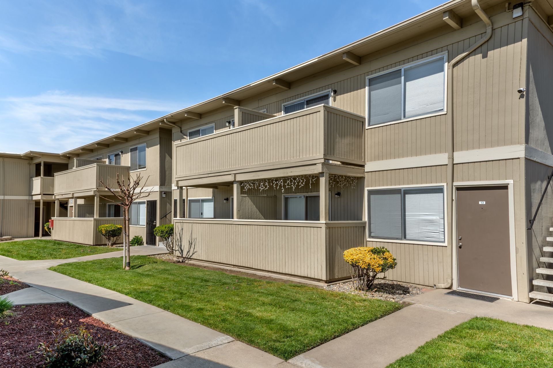 Two-story tan apartment building with balconies and a grassy yard under a blue sky.