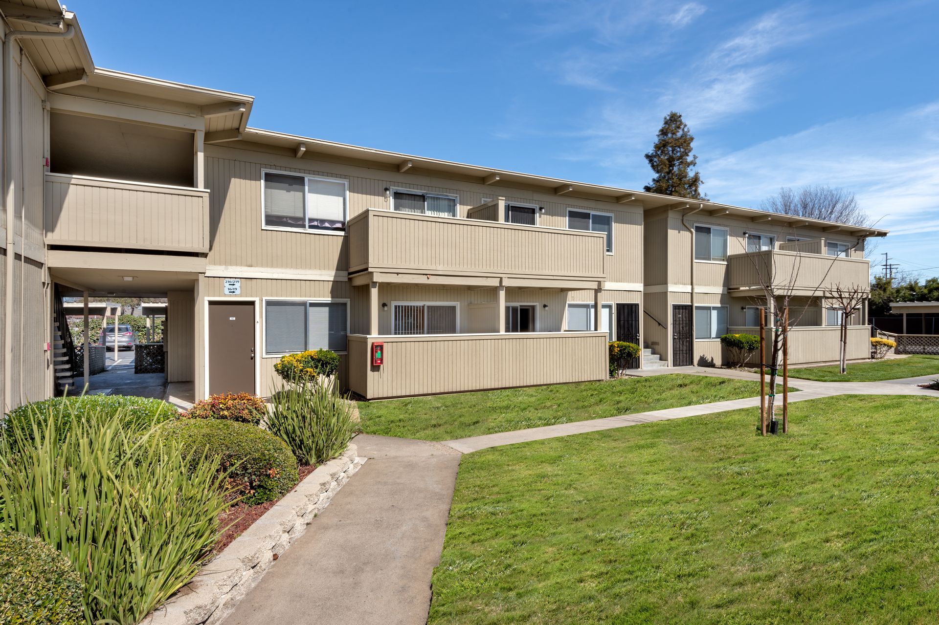Apartment building with tan siding, balconies, and a grassy lawn on a sunny day.