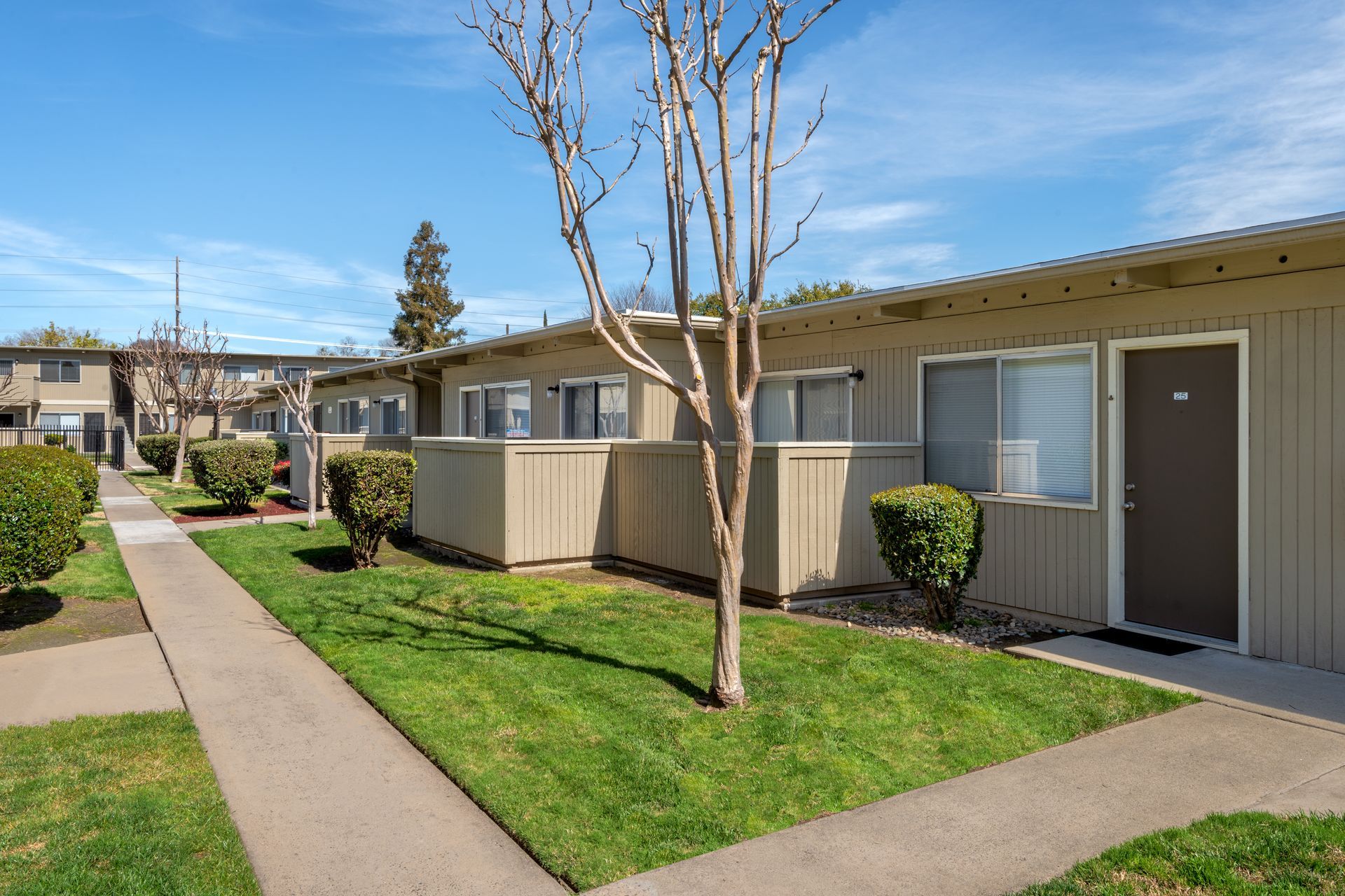 Apartment buildings with tan exteriors, green lawns, and a sidewalk under a blue sky.
