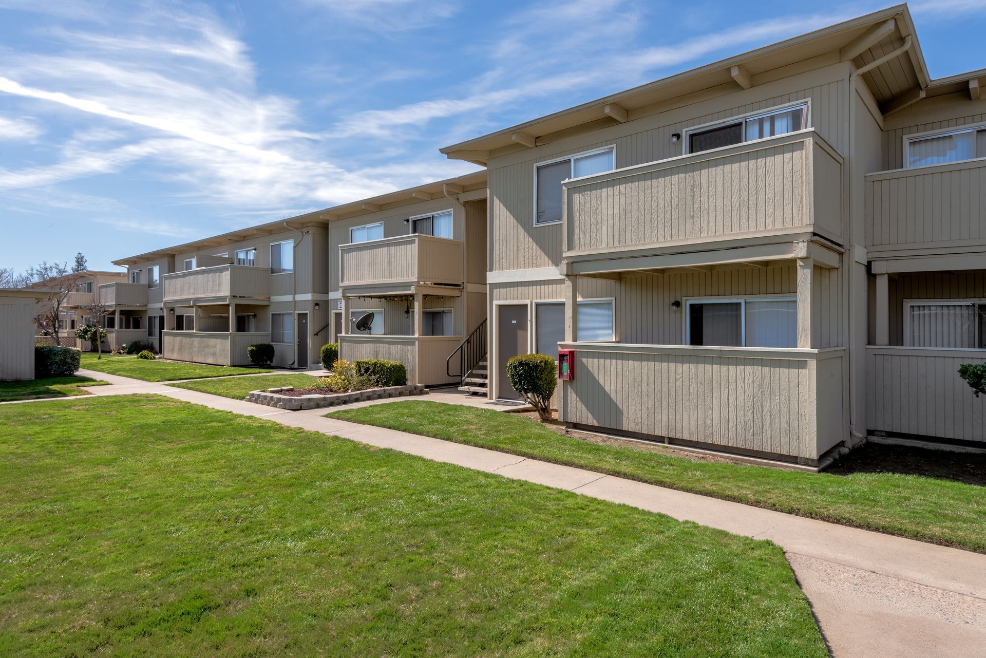 Apartment complex with beige buildings, balconies, green grass, and a sunny blue sky.