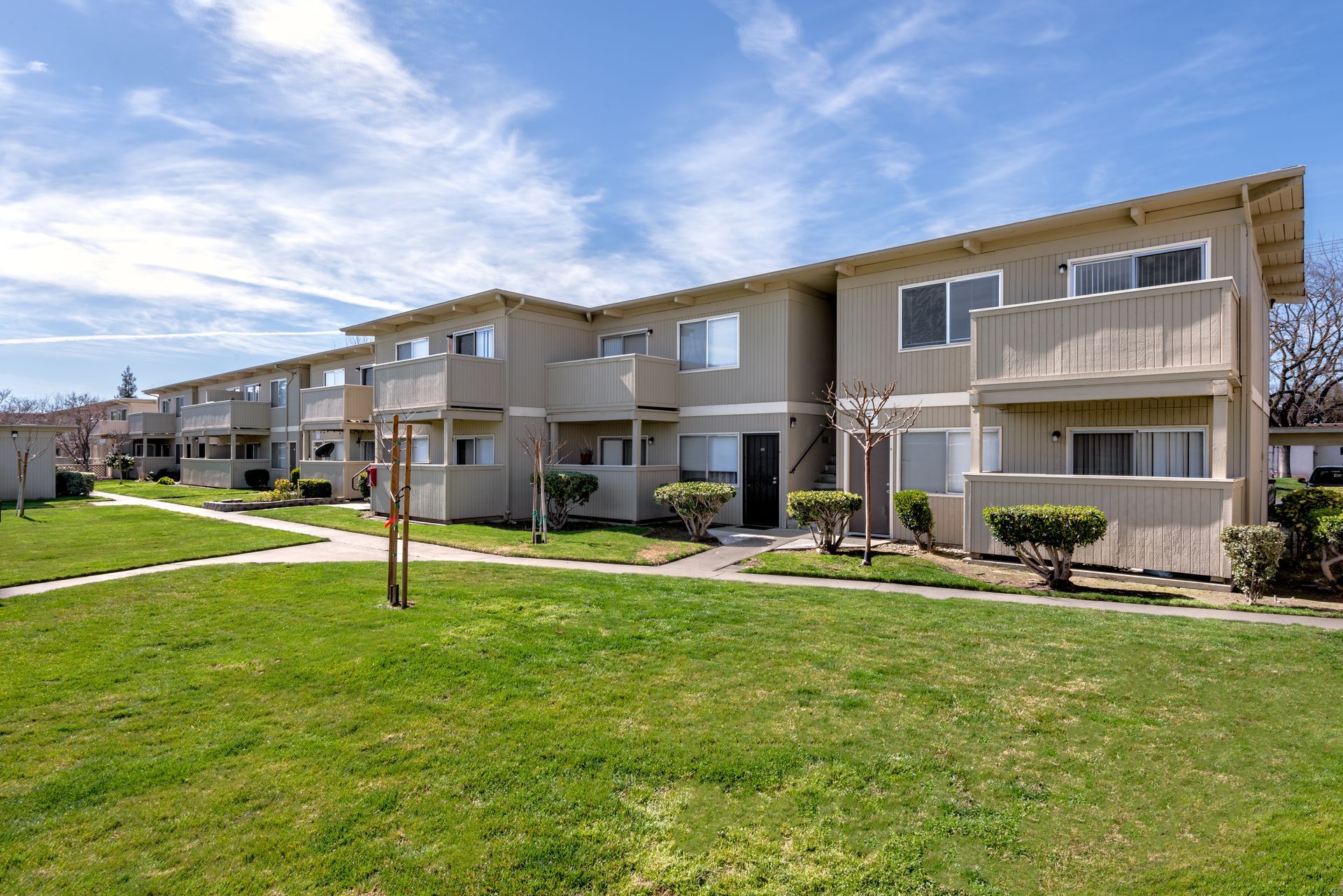 Two-story apartment building with beige siding, balconies, and a green lawn under a blue sky.