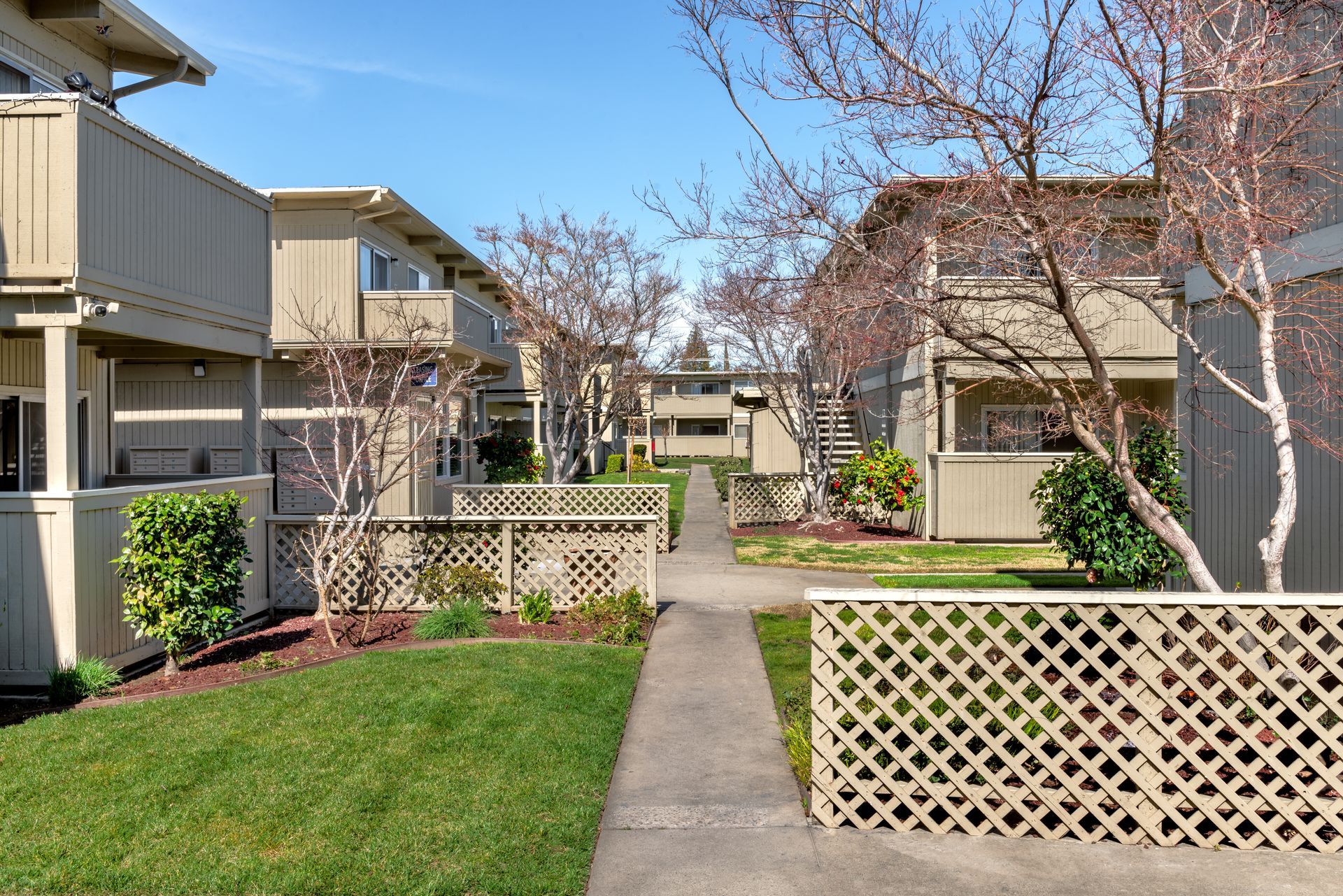 Apartment complex with beige buildings, a concrete path, and green lawns under a blue sky.