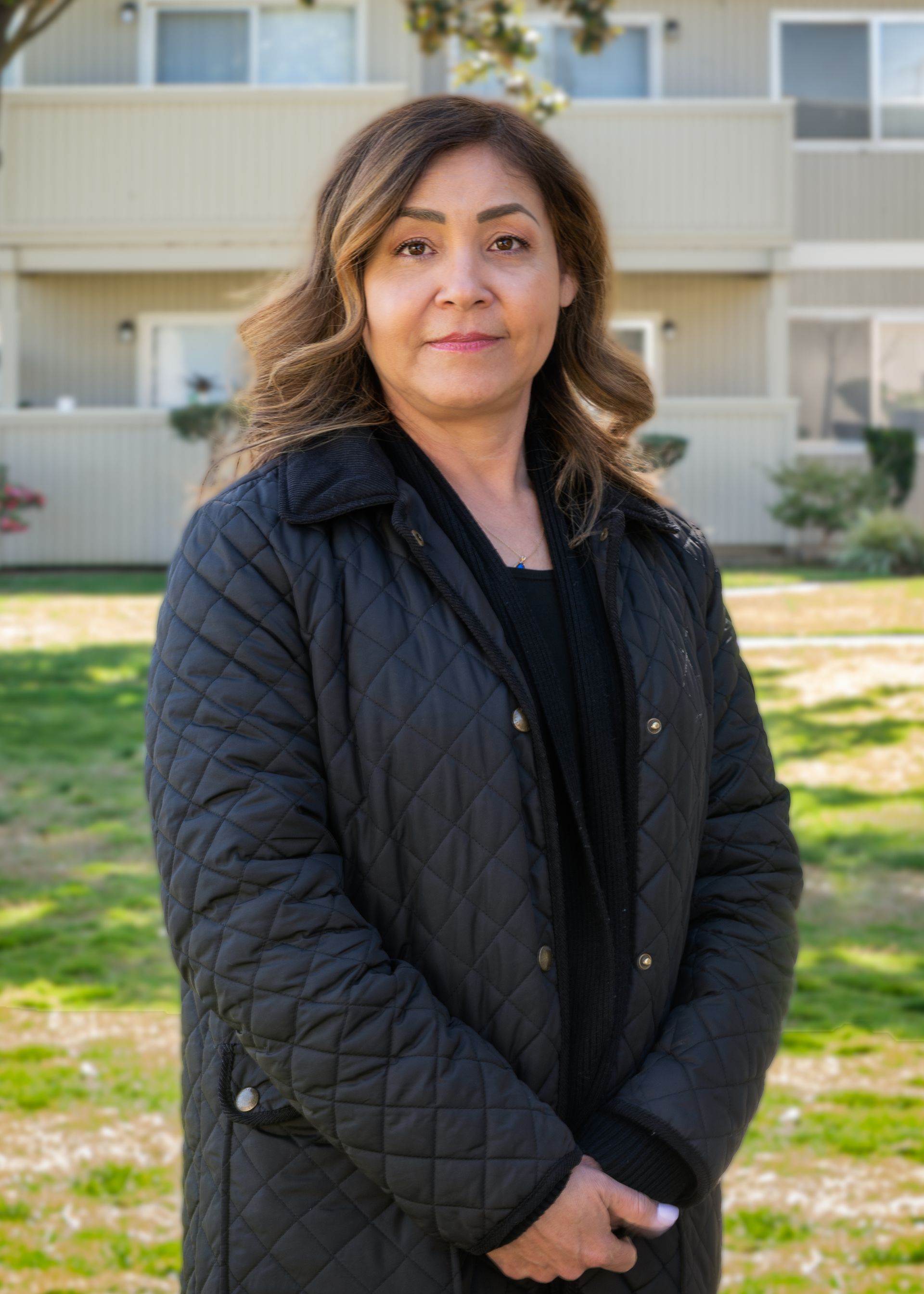 Woman standing outdoors in front of a building, wearing a black jacket, looking at the camera.