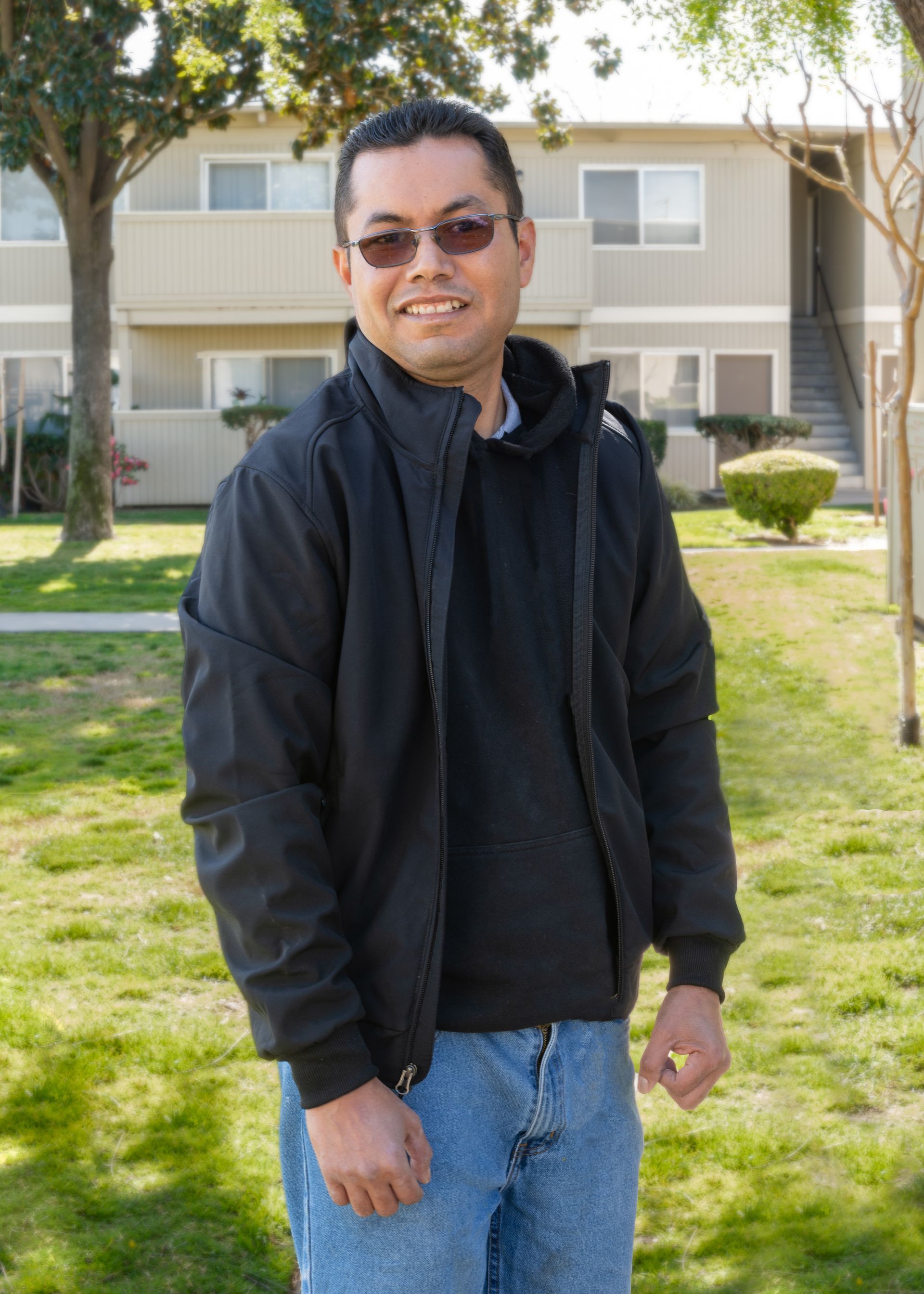 Man wearing black jacket and jeans smiles outdoors in front of a building.