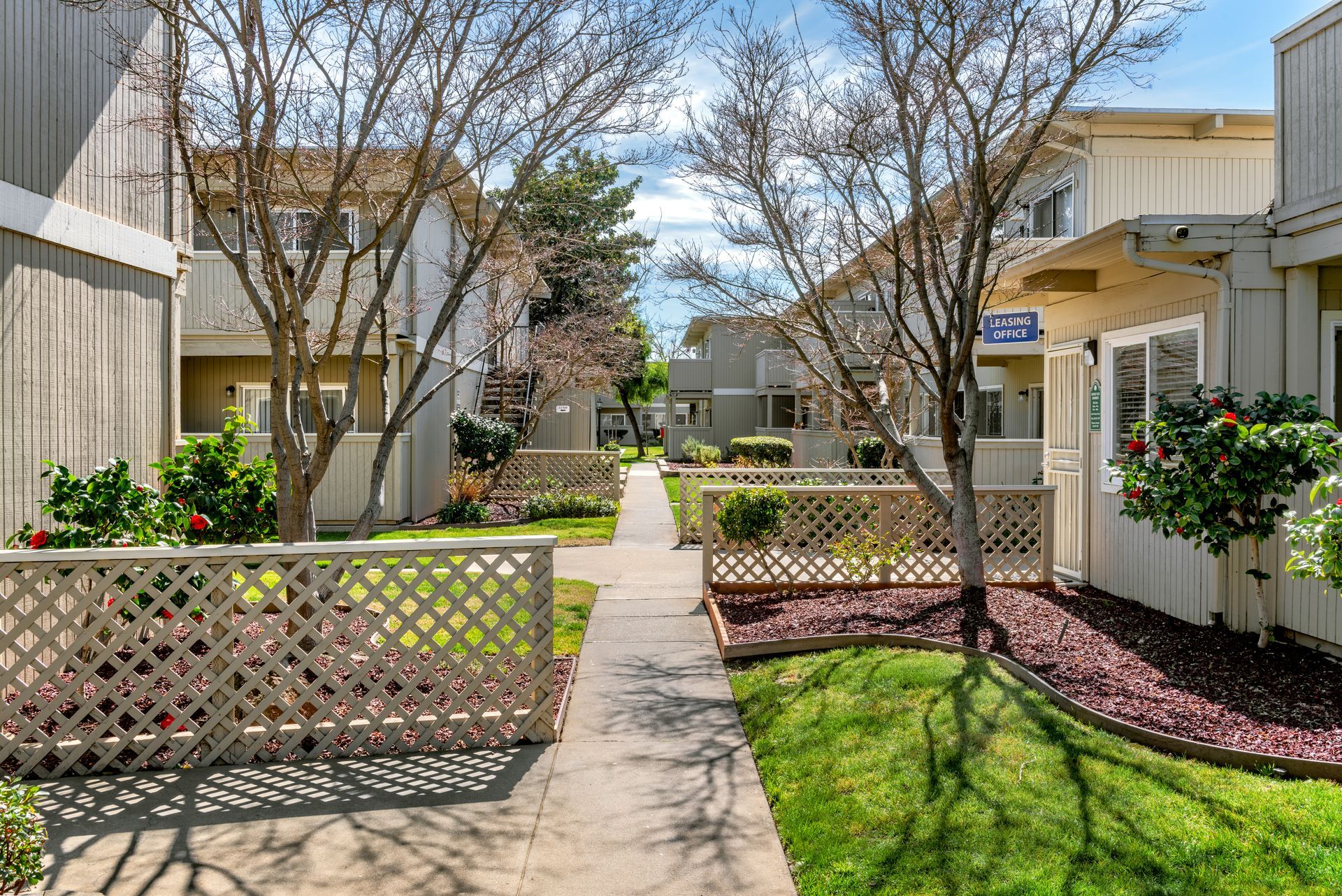 Apartment complex exterior with pathway, fences, trees, and green lawn under a partly cloudy sky.