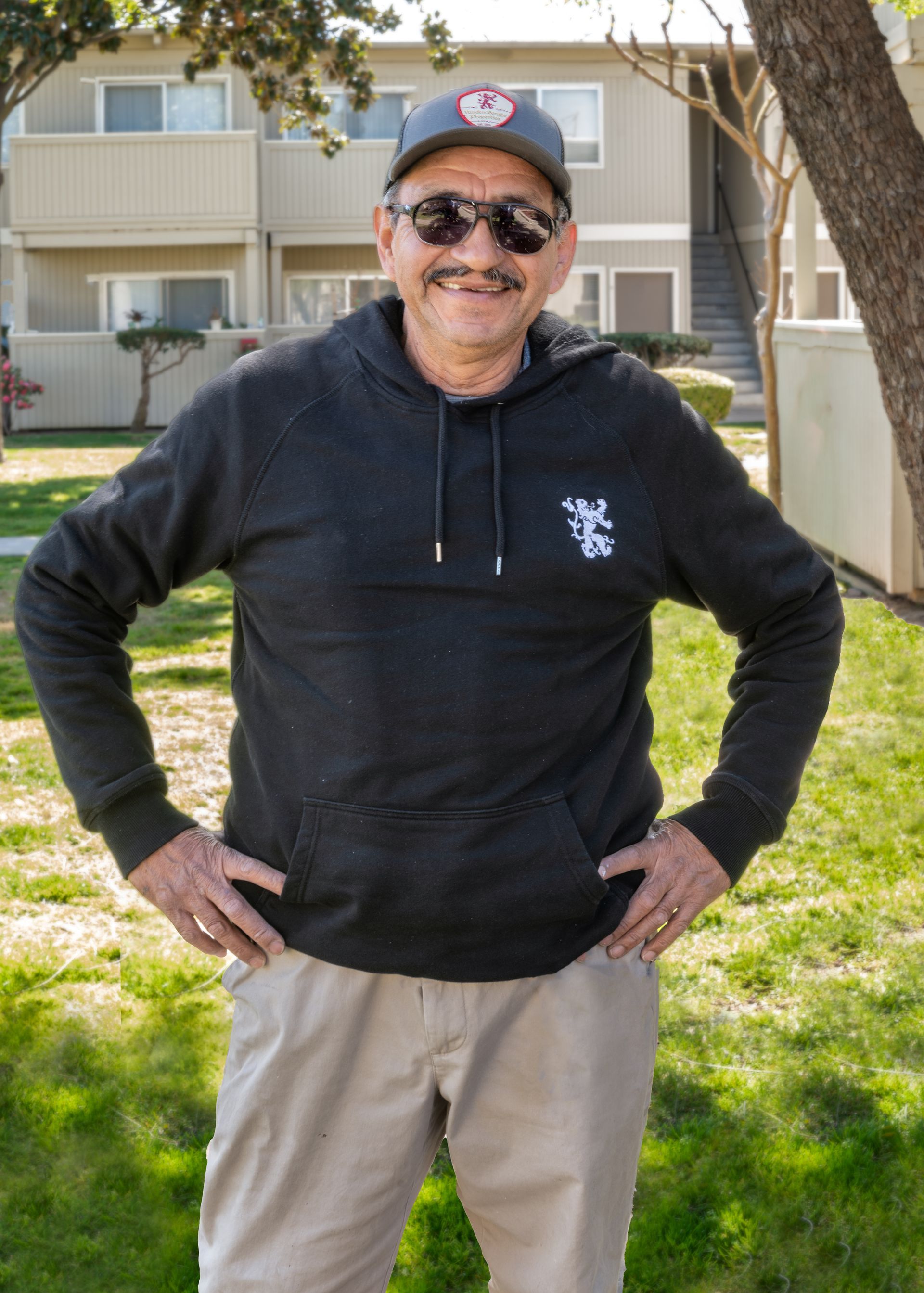 Man in gray hoodie and cap, sunglasses, hands on hips, in front of apartment building.
