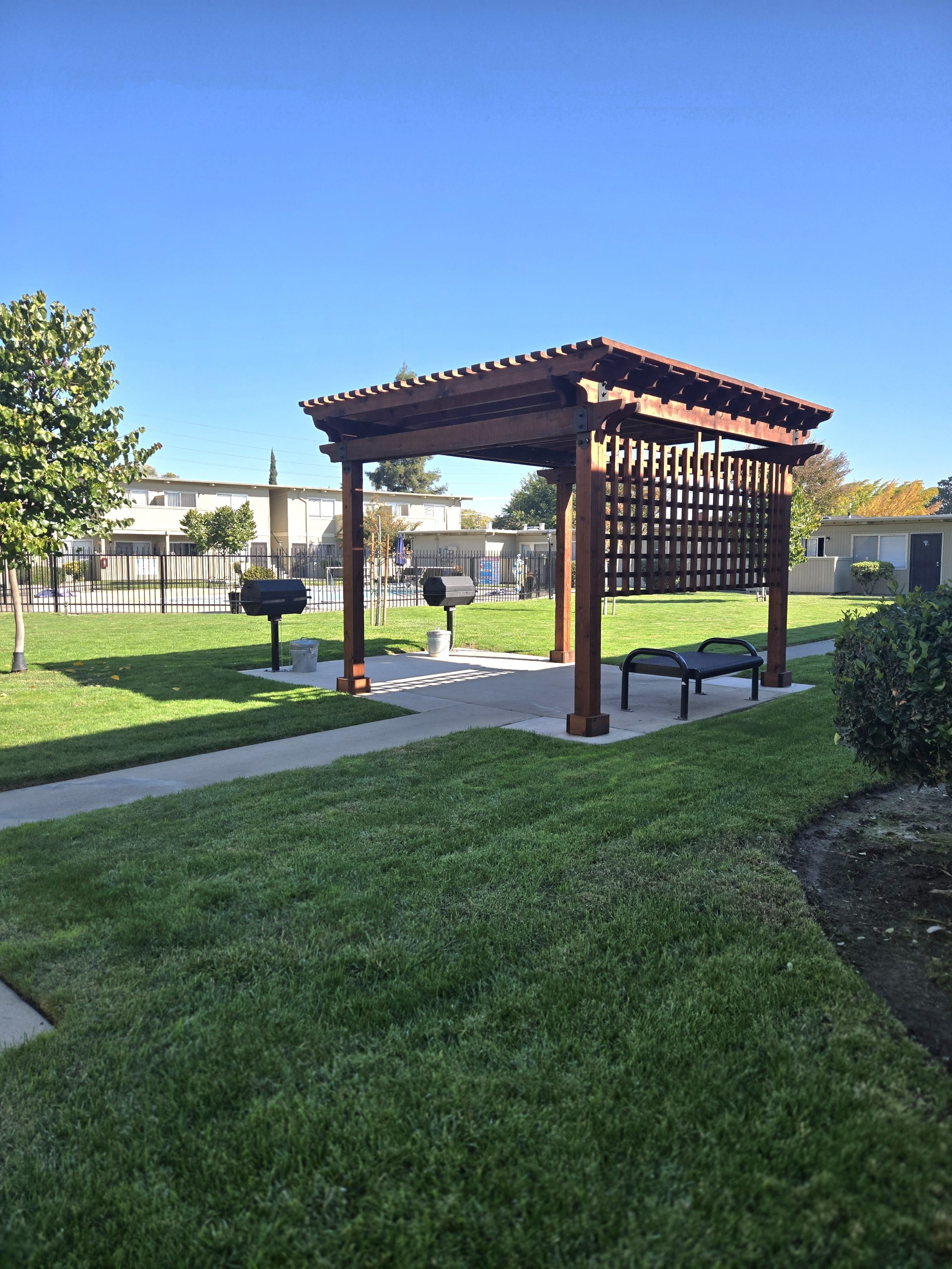 Grassy outdoor area with grills, a bench, and a pool behind a fence, in front of apartments.