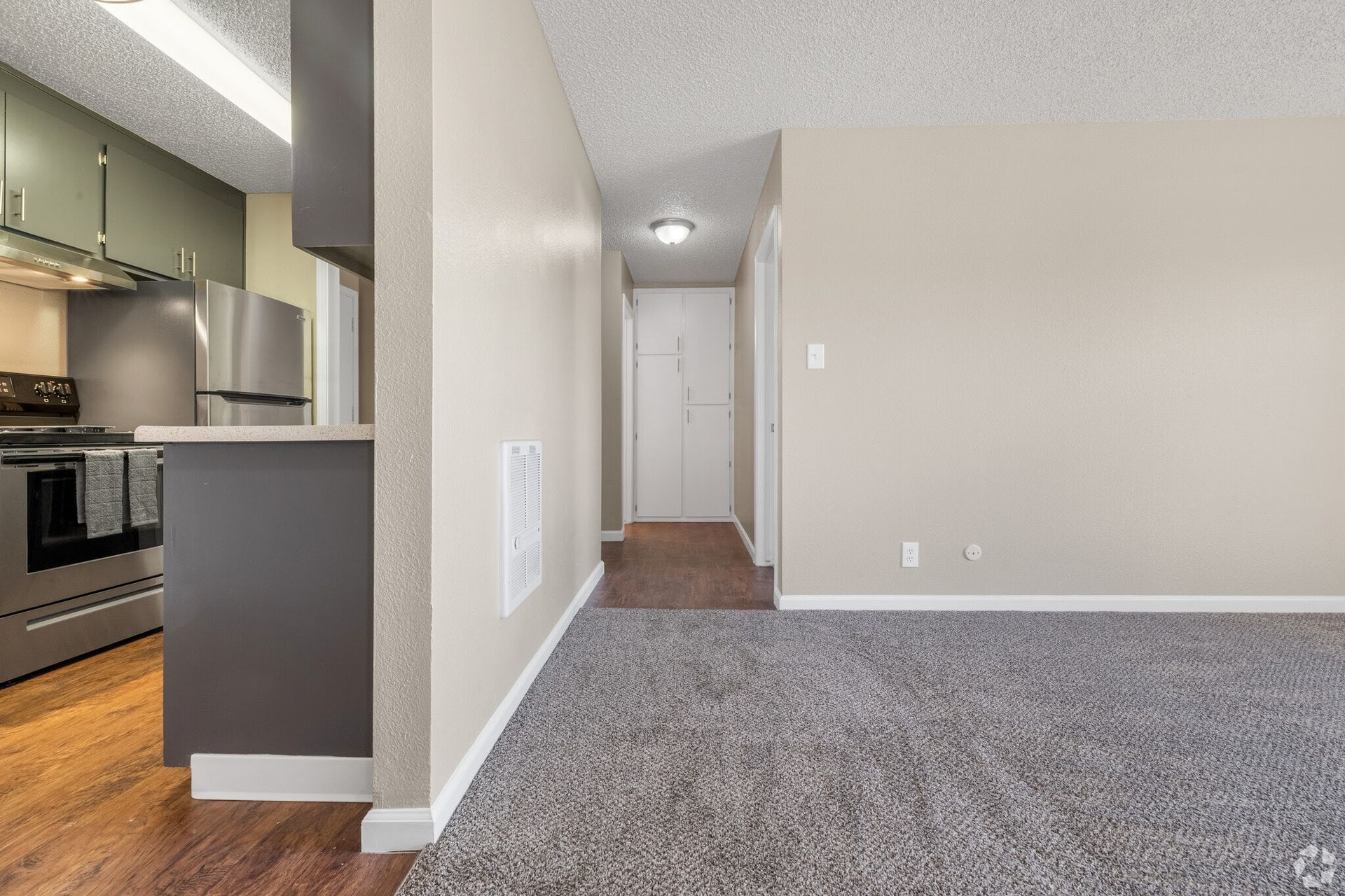 Apartment interior view: kitchen with stainless steel appliances, hallway with white door, gray carpet.