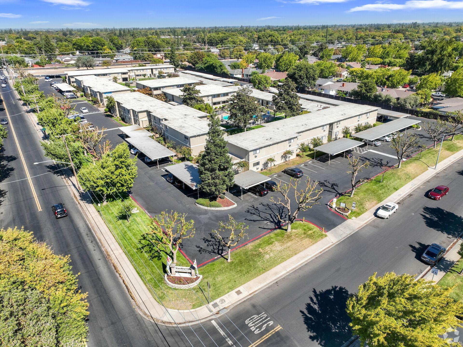 Aerial view of an apartment complex with multiple buildings, carports, and a swimming pool. Green trees surround the area.
