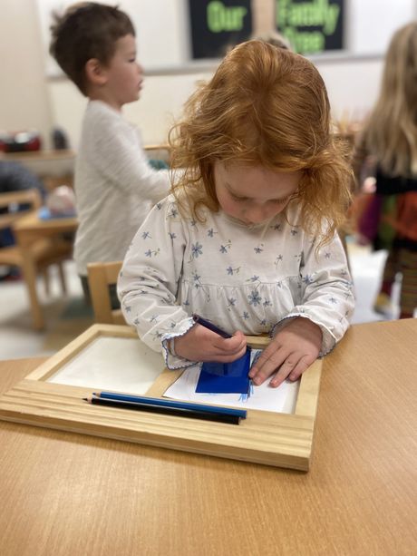 A child is drawing on a piece of paper with a blue pencil using Montessori materials.