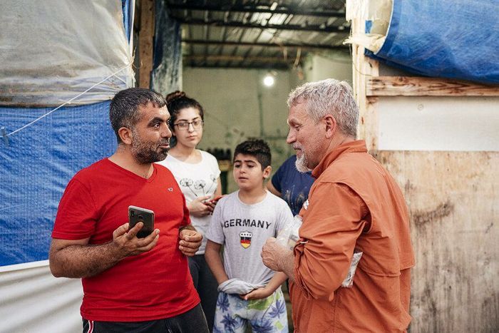 Two men converse outside a makeshift shelter, with a woman and boy standing nearby.