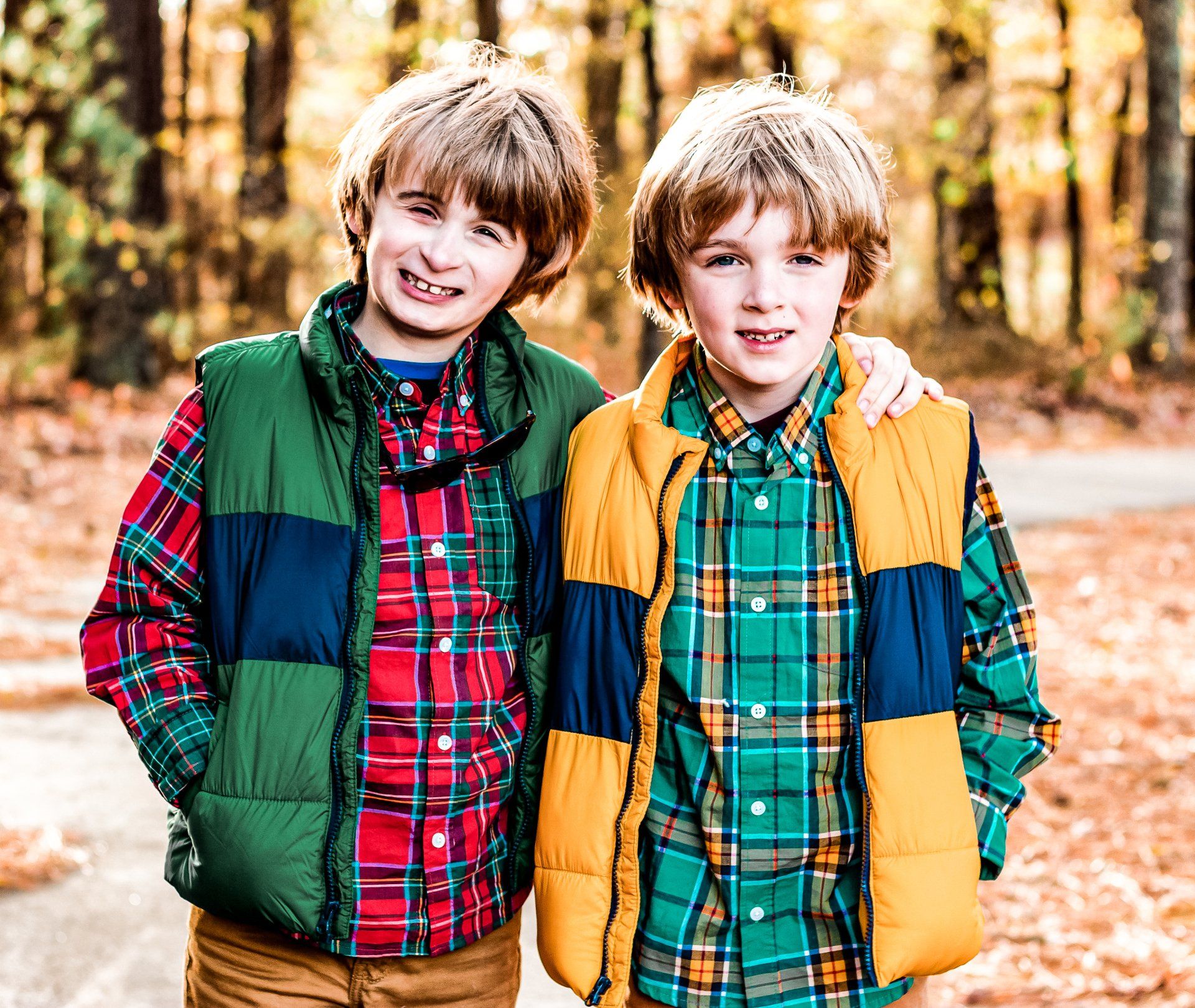 Two young boys wearing plaid shirts and vests are standing next to each other in the woods.