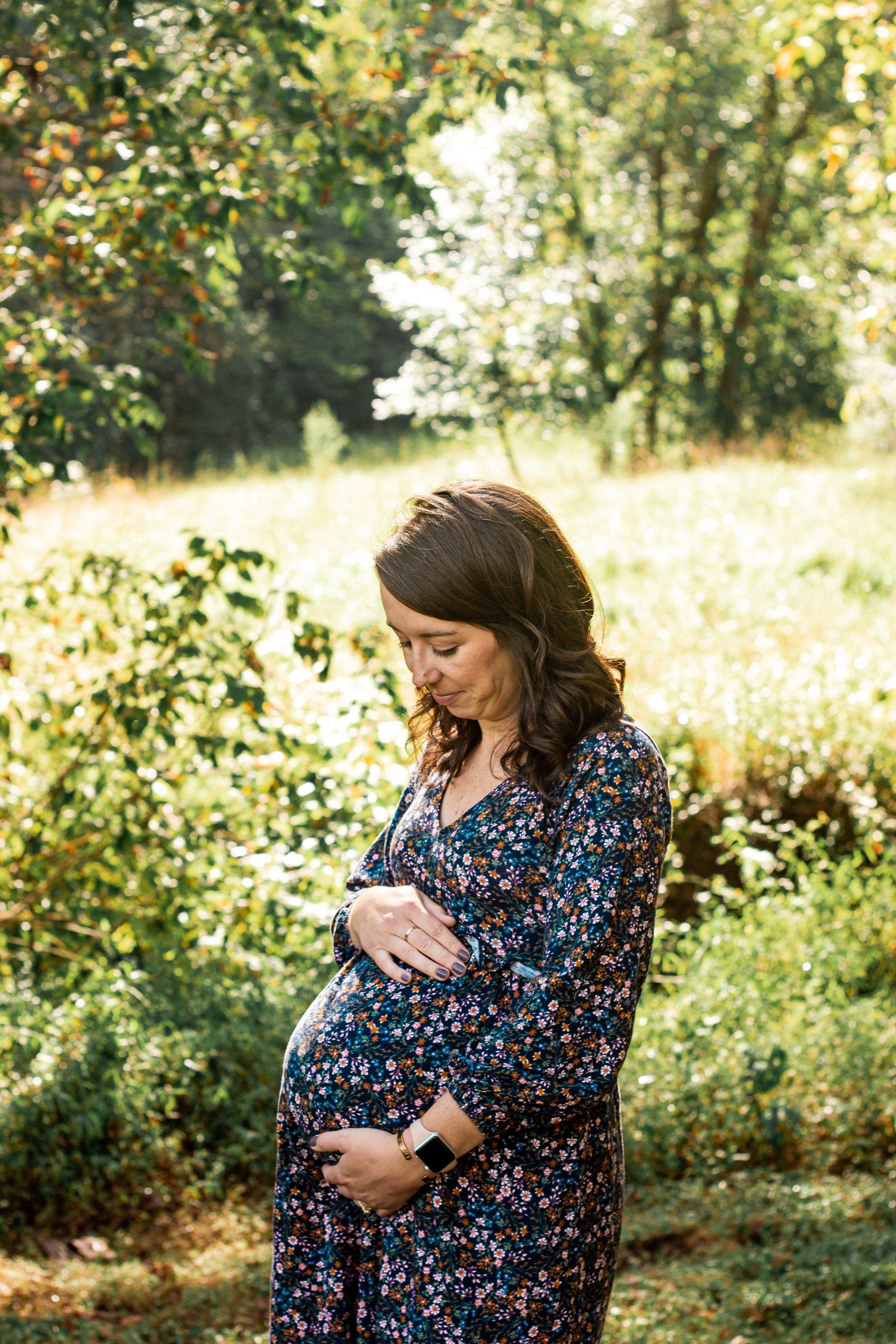 A pregnant woman in a blue dress is standing in a field holding her belly.