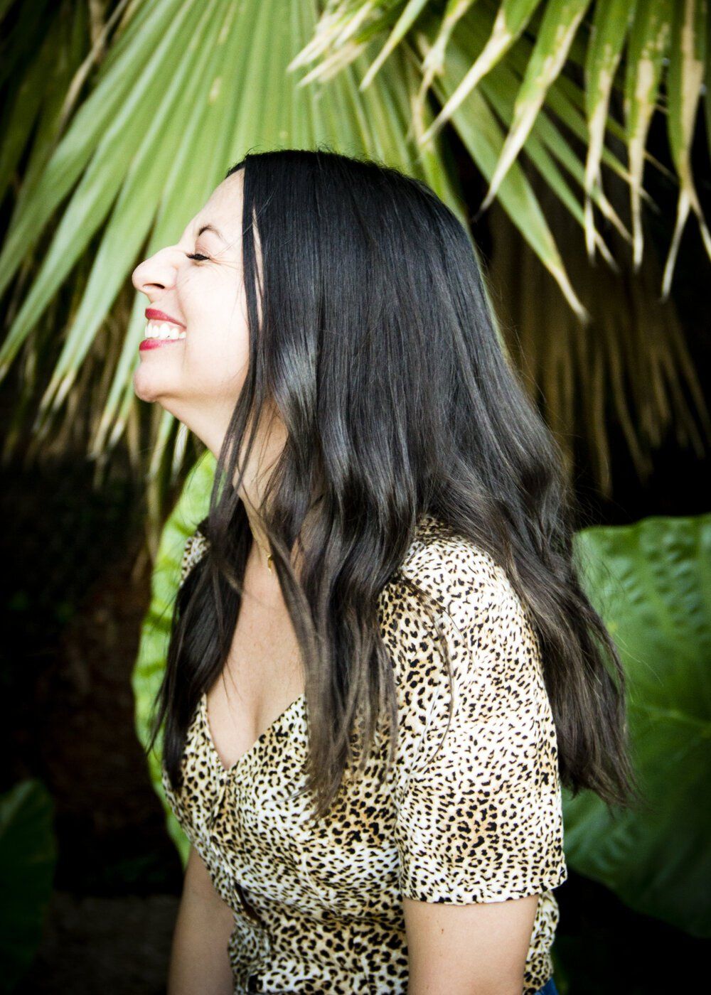 A woman in a leopard print top is smiling in front of a palm tree.