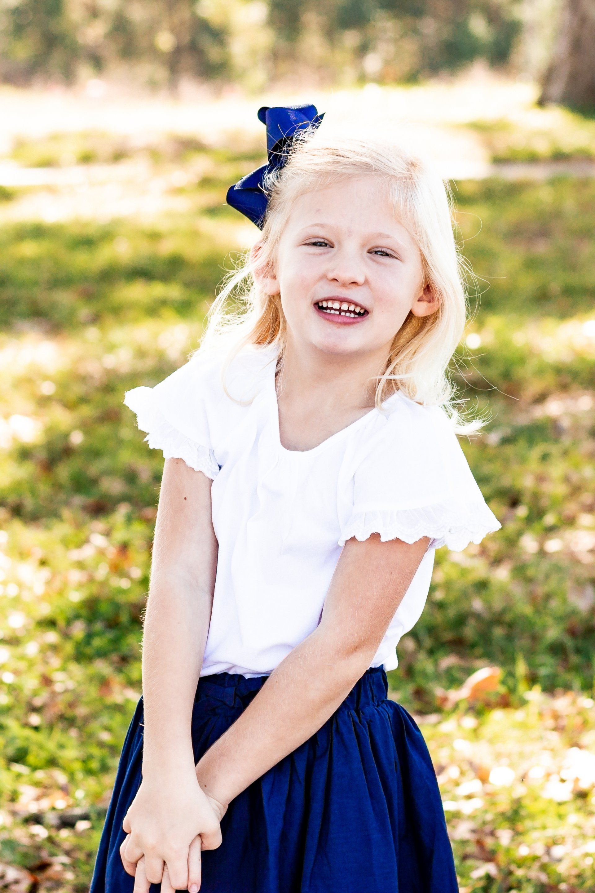 A little girl wearing a white shirt and a blue skirt is standing in the grass.