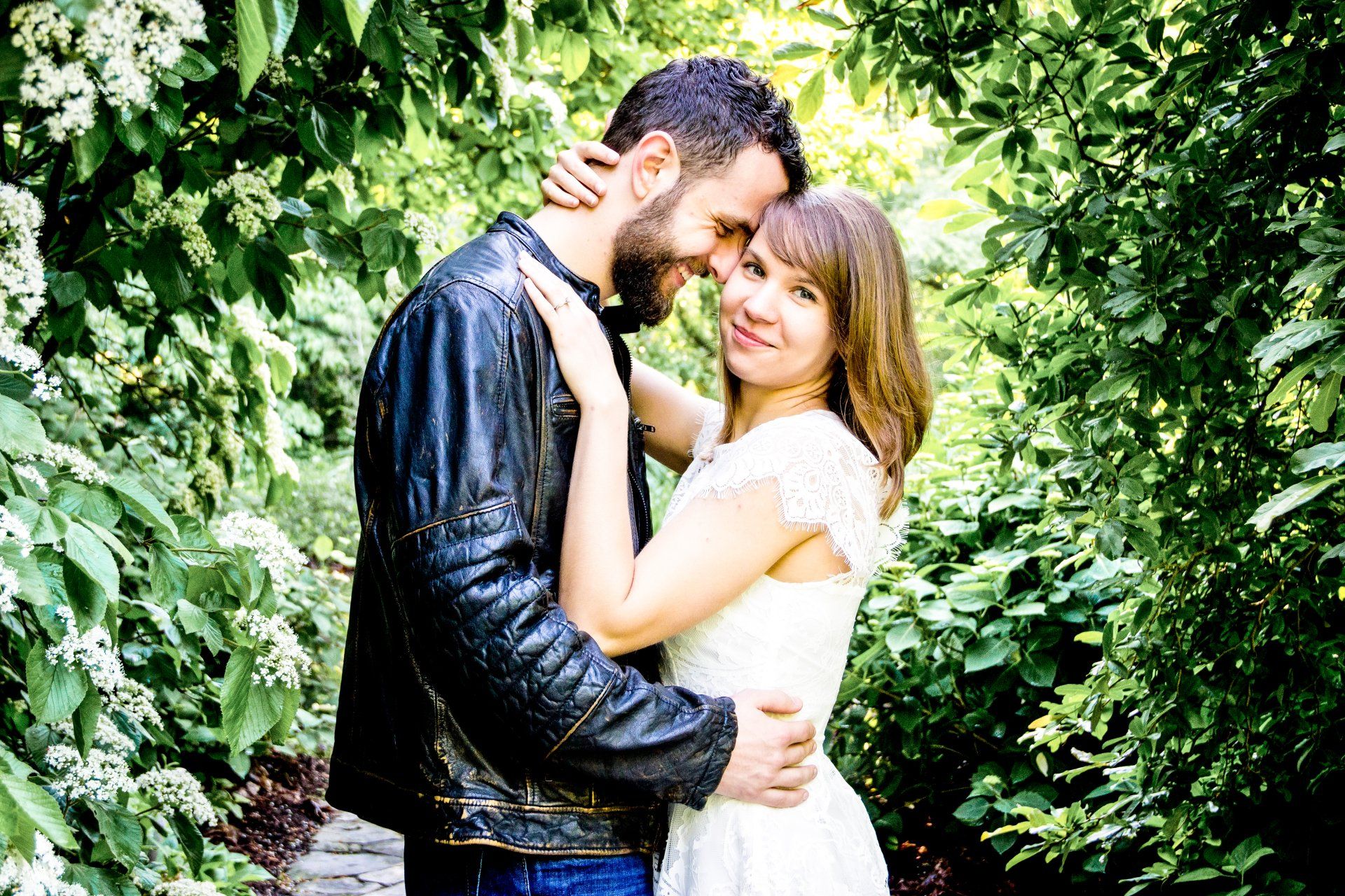 A man in a leather jacket is hugging a woman in a white dress.