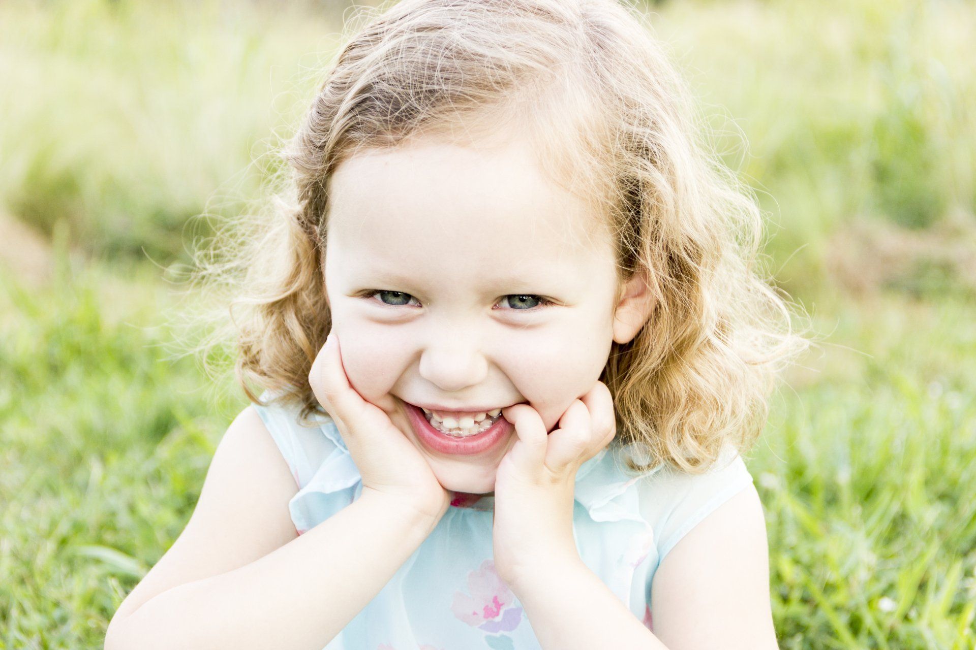 A little girl is sitting in the grass with her hands on her face and smiling.