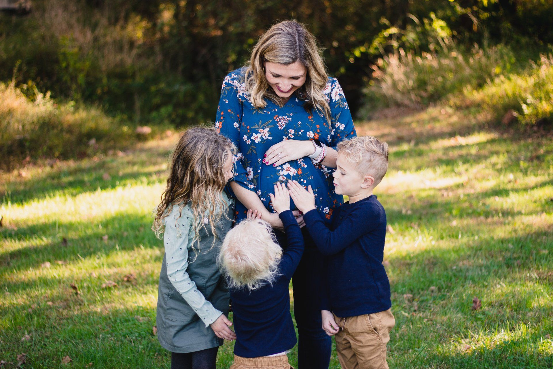 A pregnant woman is surrounded by three children in a park.