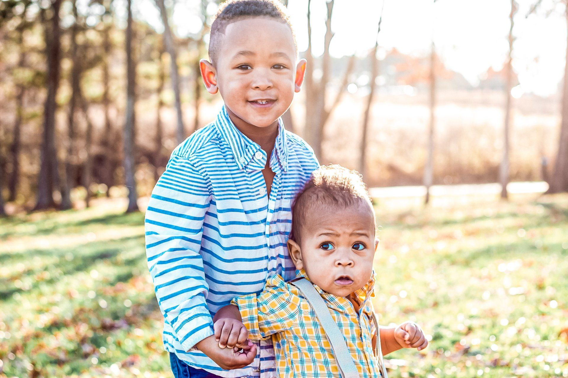 Two young boys are standing next to each other in a park holding hands.