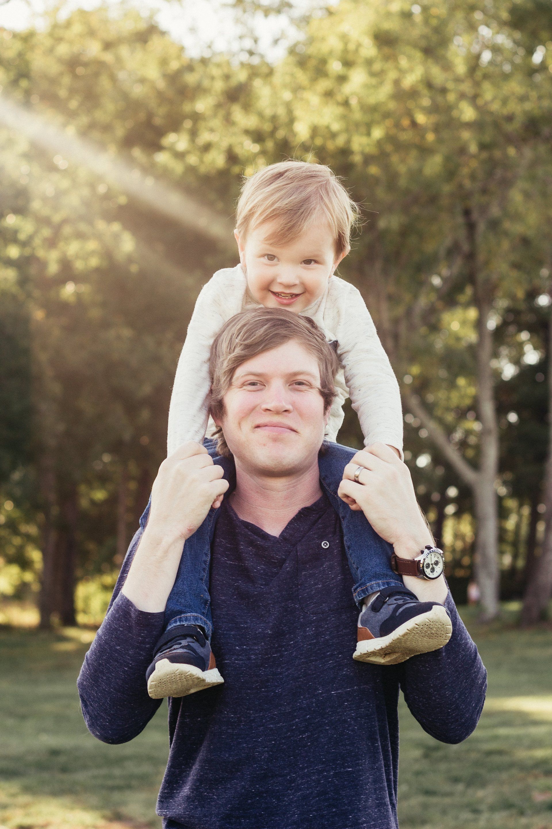 A man is carrying a little boy on his shoulders in a park.