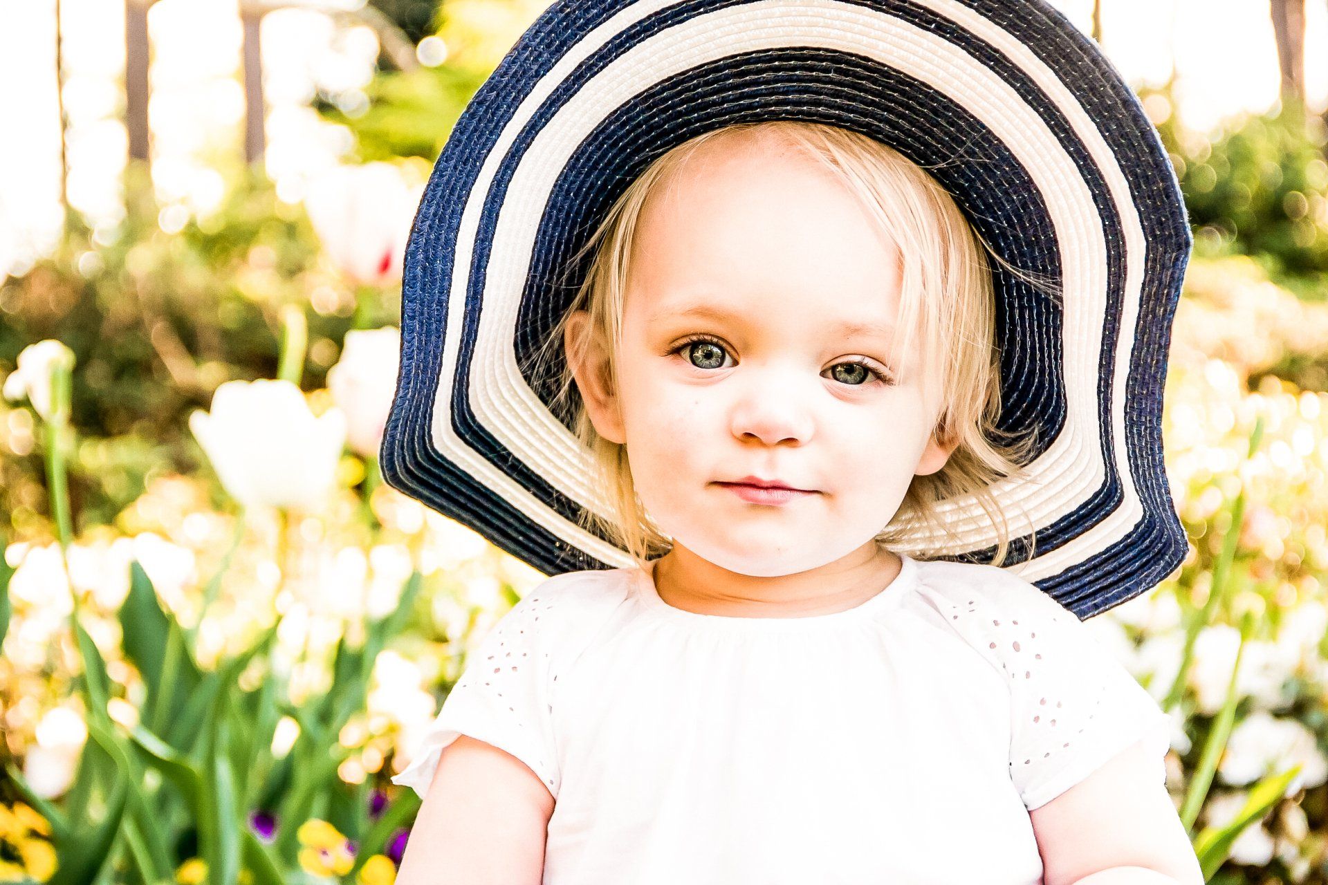 A little girl wearing a hat is sitting in a field of flowers.
