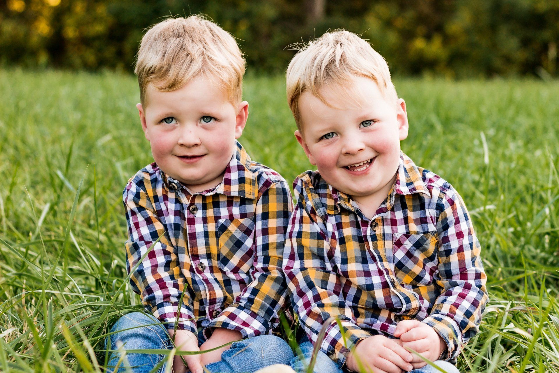 Two young boys in plaid shirts are sitting next to each other in the grass.