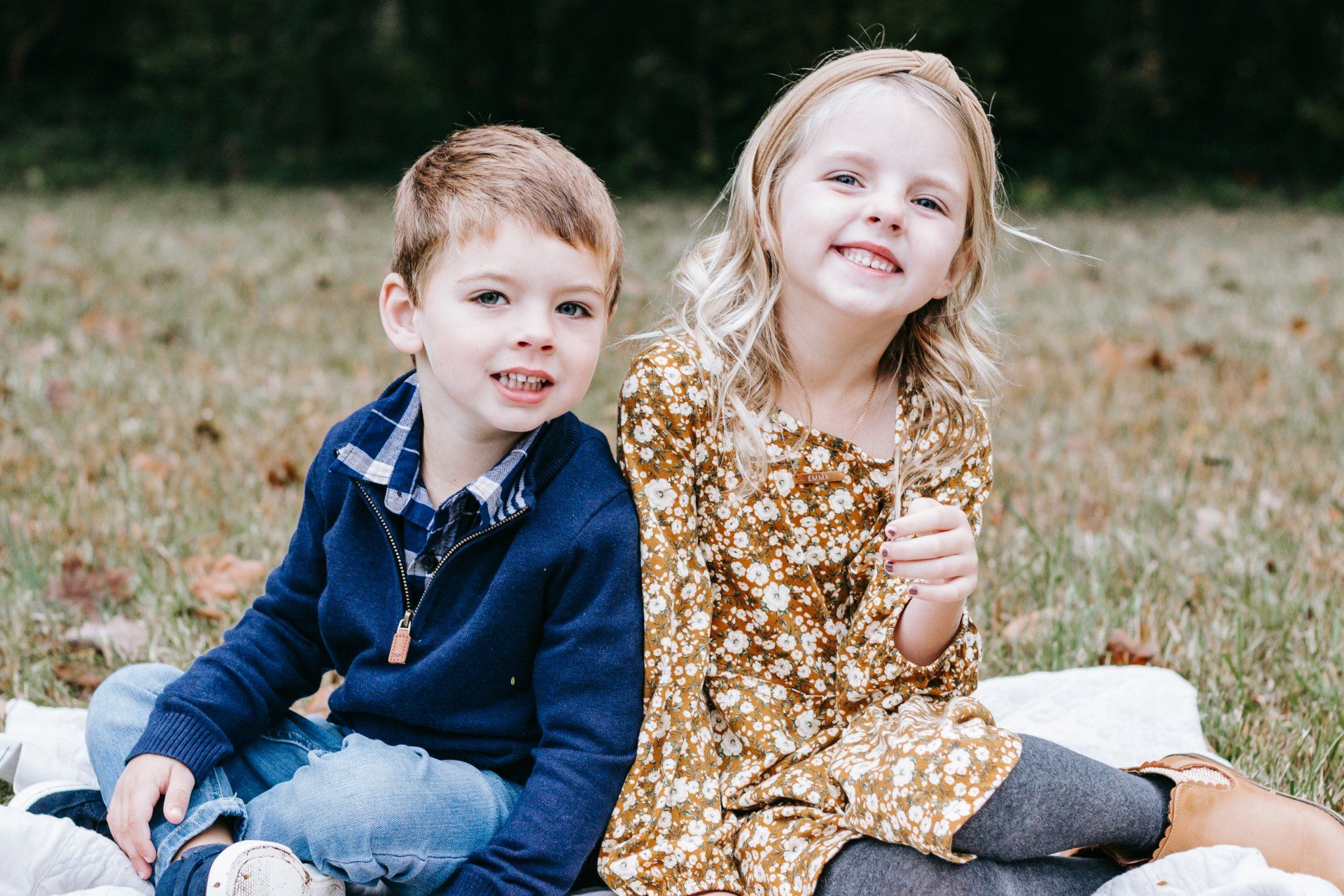 A boy and a girl are sitting on a blanket in the grass.