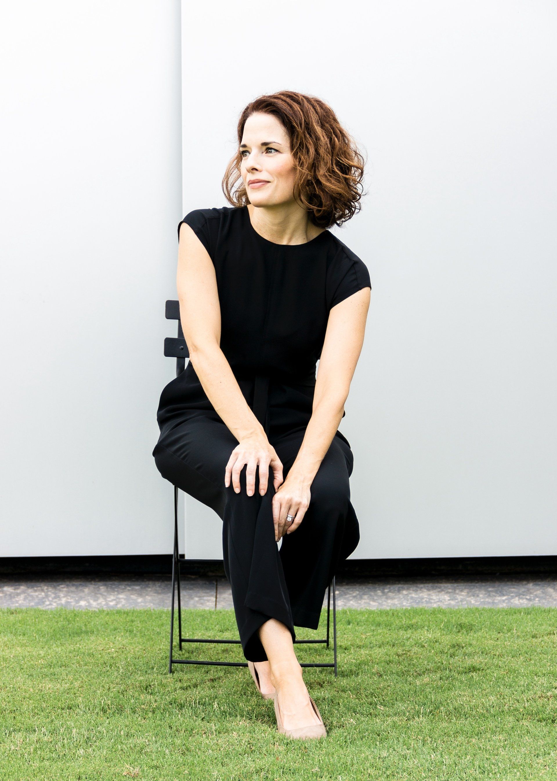 A woman in a black jumpsuit is sitting on a chair in the grass.