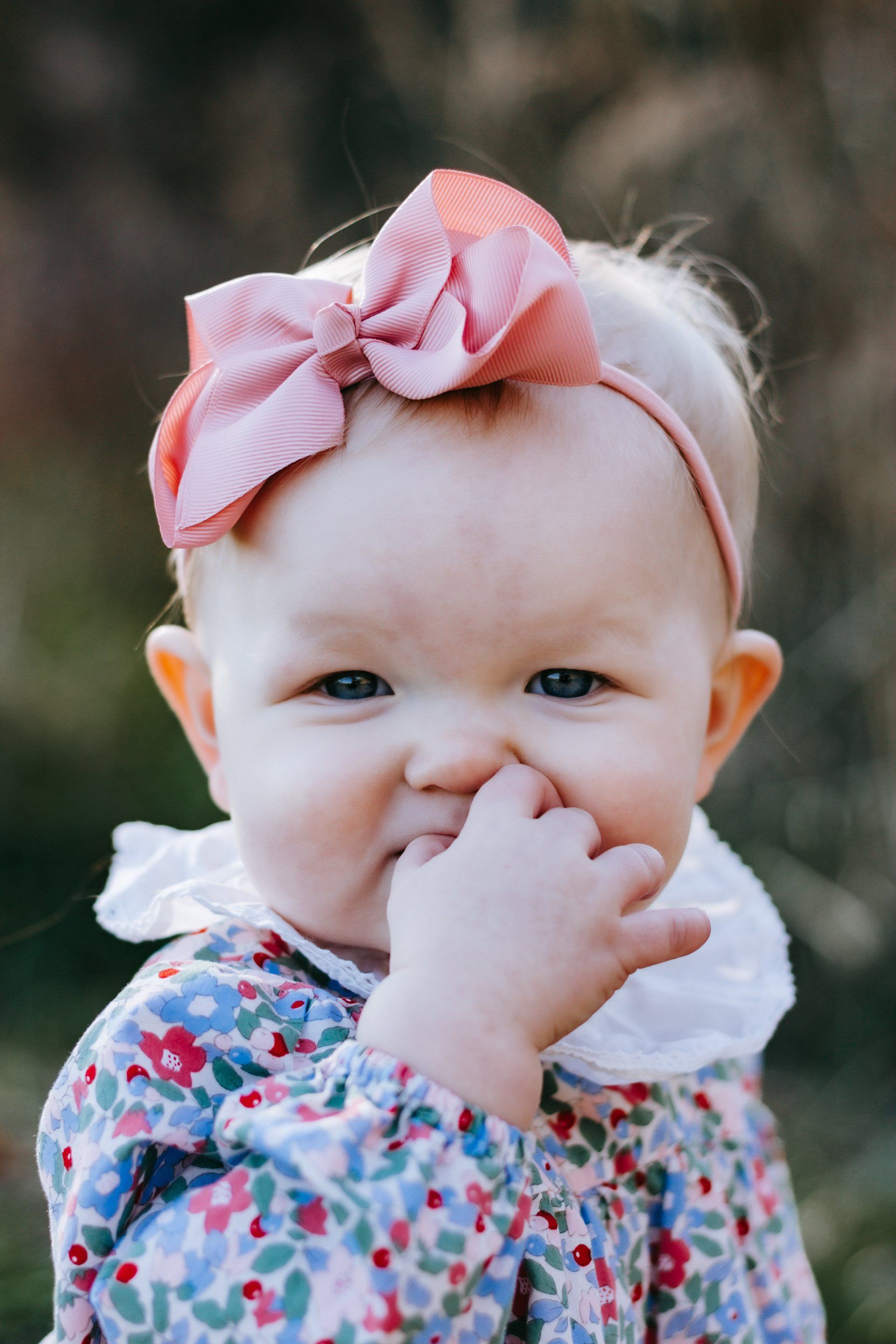 A baby with a pink bow on her head is covering her nose with her hand.