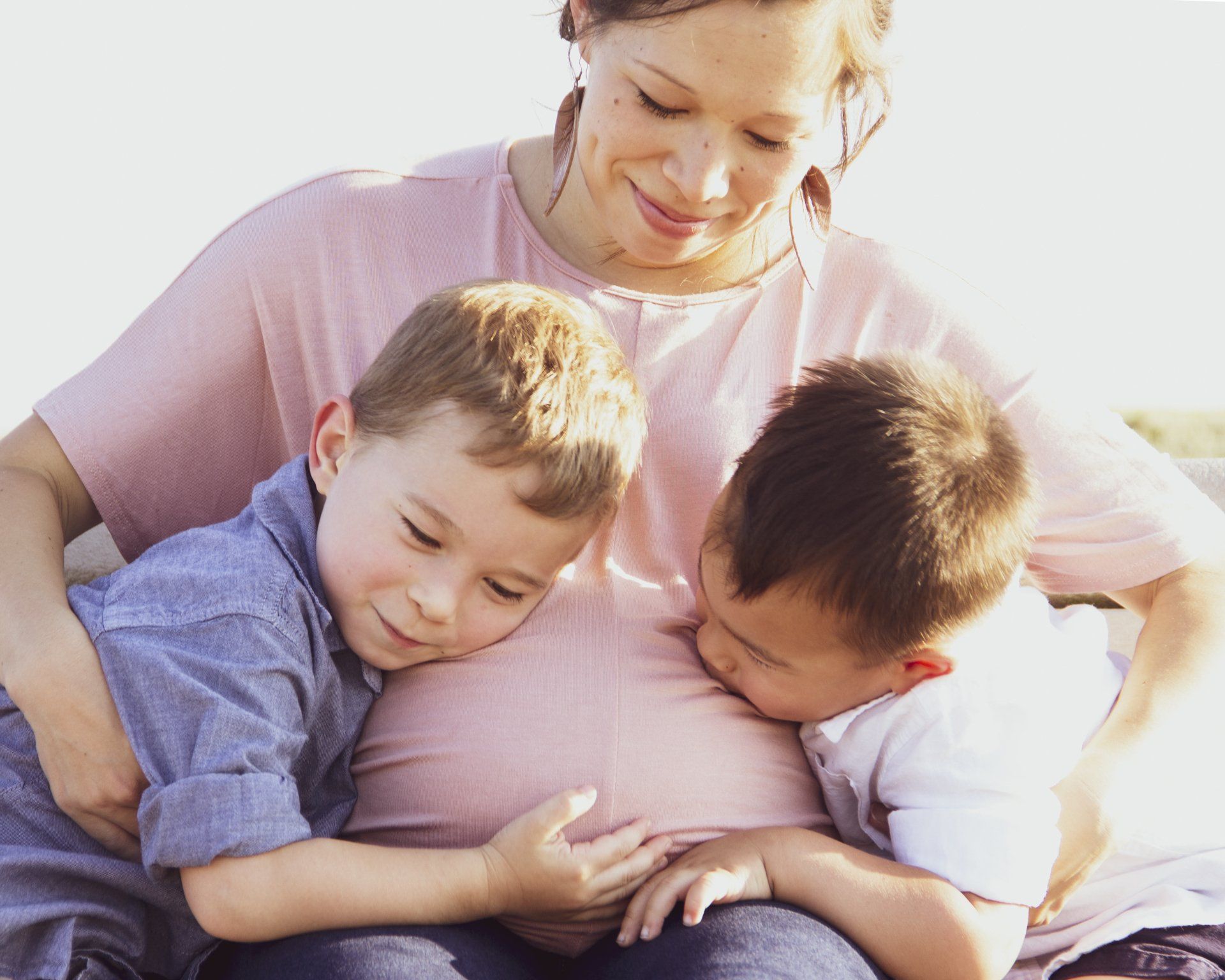 A pregnant woman is sitting with two boys hugging her belly.