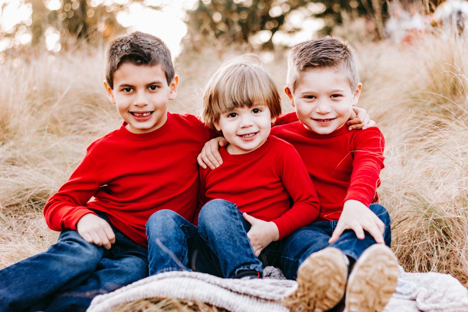 Three young boys in red shirts are sitting on a blanket in a field.