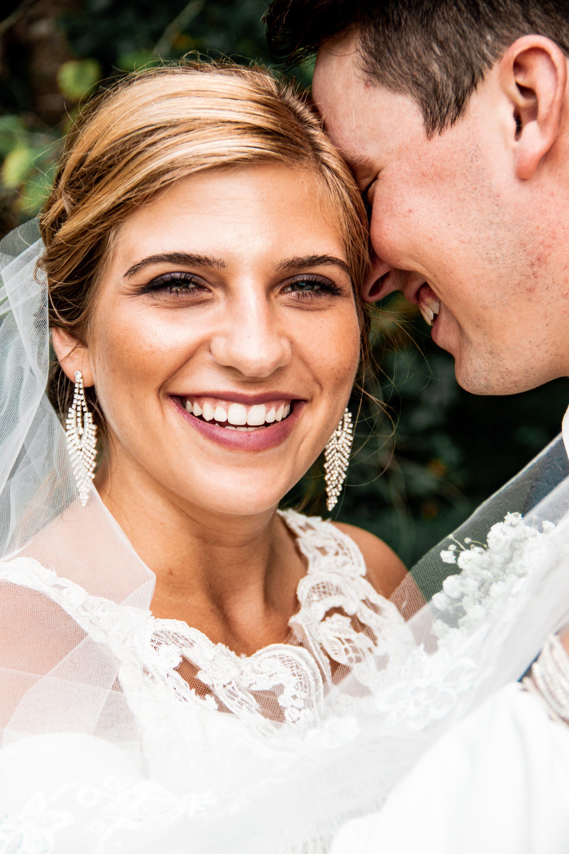 A bride and groom are posing for a picture and the bride is smiling while the groom kisses her forehead.