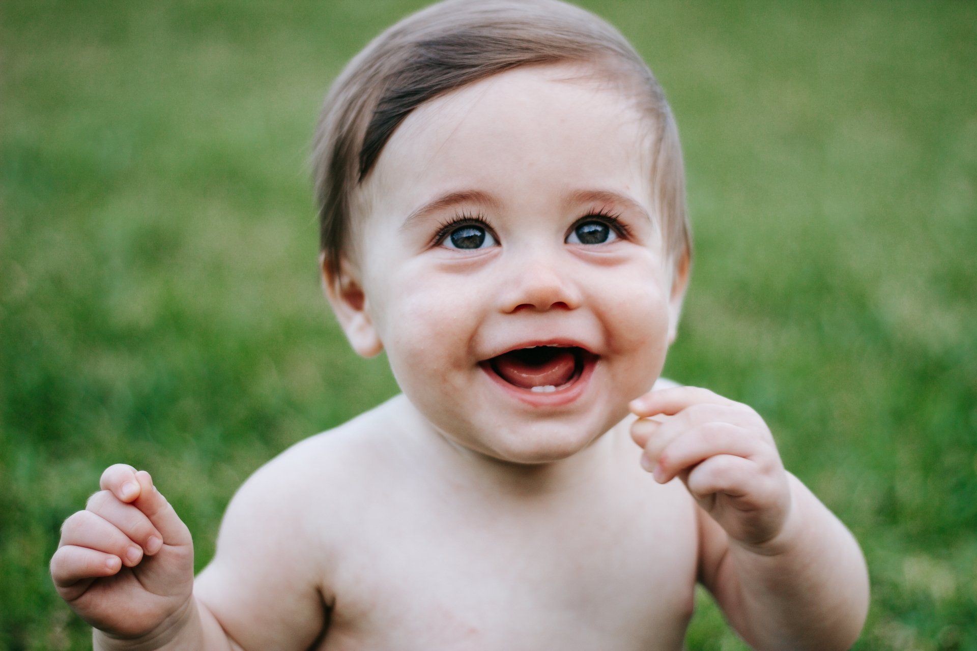 A baby without a shirt is sitting in the grass and smiling.