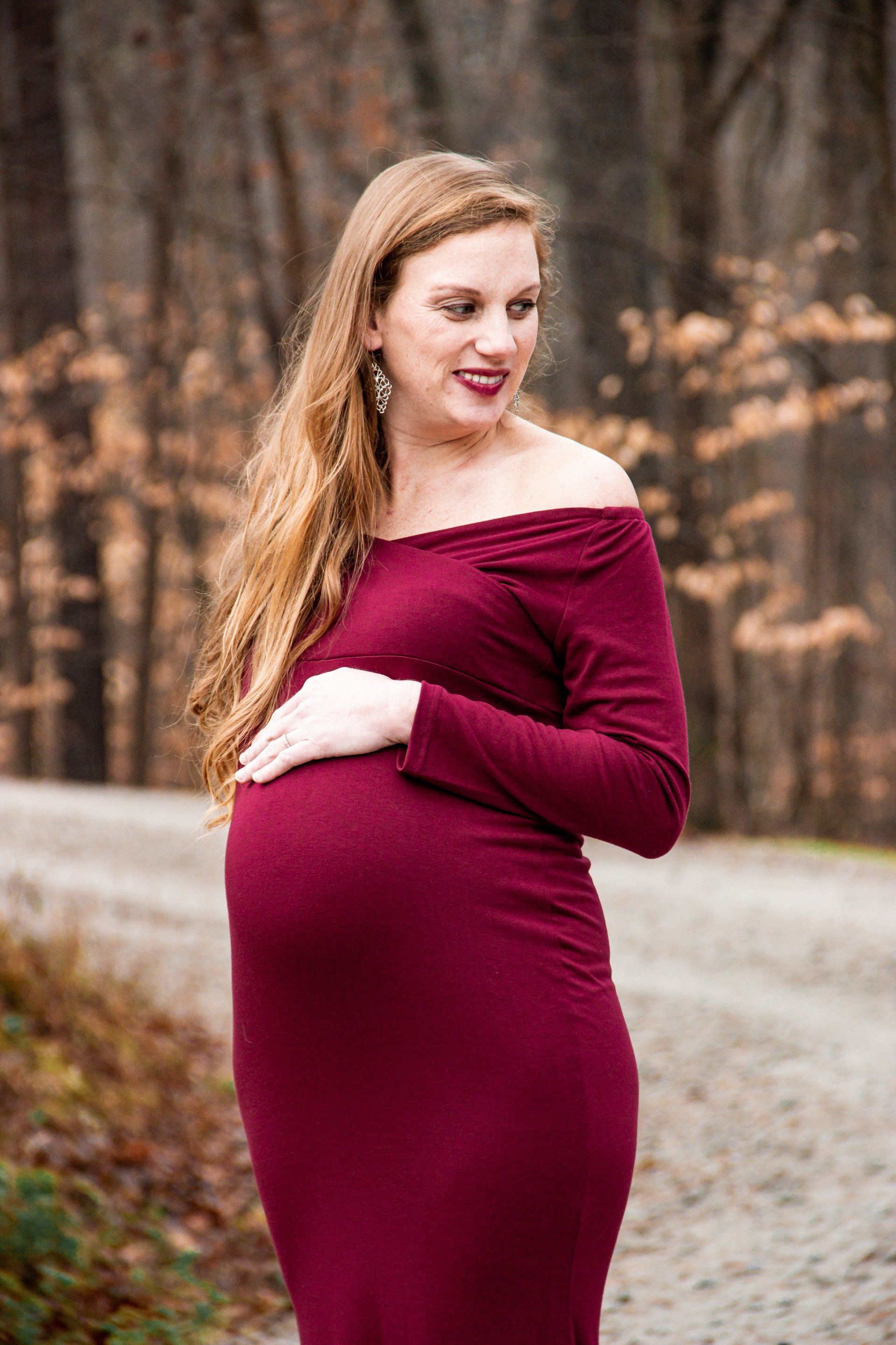 A pregnant woman in a red dress is standing on a dirt road holding her belly.